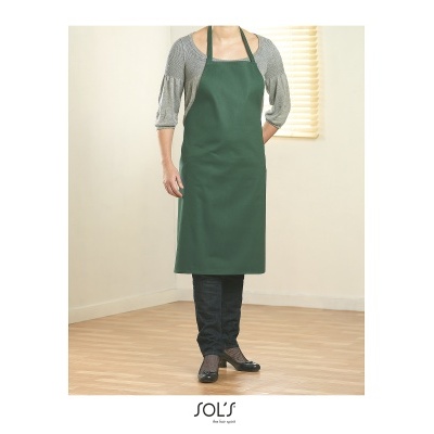 Pessoa com avental verde escuro sobre blusa cinza, em ambiente interno com parede clara e piso de madeira.