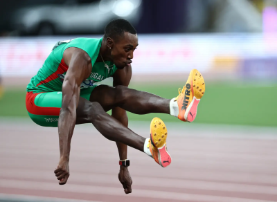 Atleta a saltar em competição de atletismo com uniforme verde e sapatos amarelos.