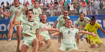Jogadoras de futebol de praia portuguesas celebrando na areia com público ao fundo.