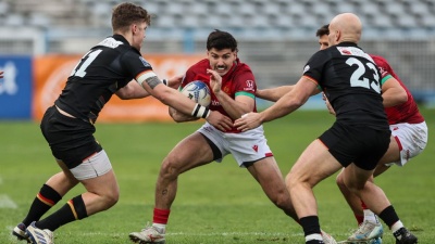 Jogadores de râguebi em campo durante jogo, um com bola e dois a defender.