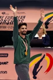 Homem com medalha dourada e casaco verde levanta os braços em competição europeia.