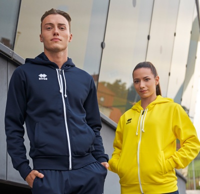 Homem com fato de treino azul escuro e mulher com fato de treino amarelo com capuz, junto a edifício com vidro refletor