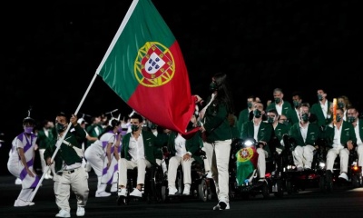 Pessoa a portar a bandeira de Portugal durante evento noturno, com pessoas vestidas de fatos verdes e brancos sentadas ao fundo