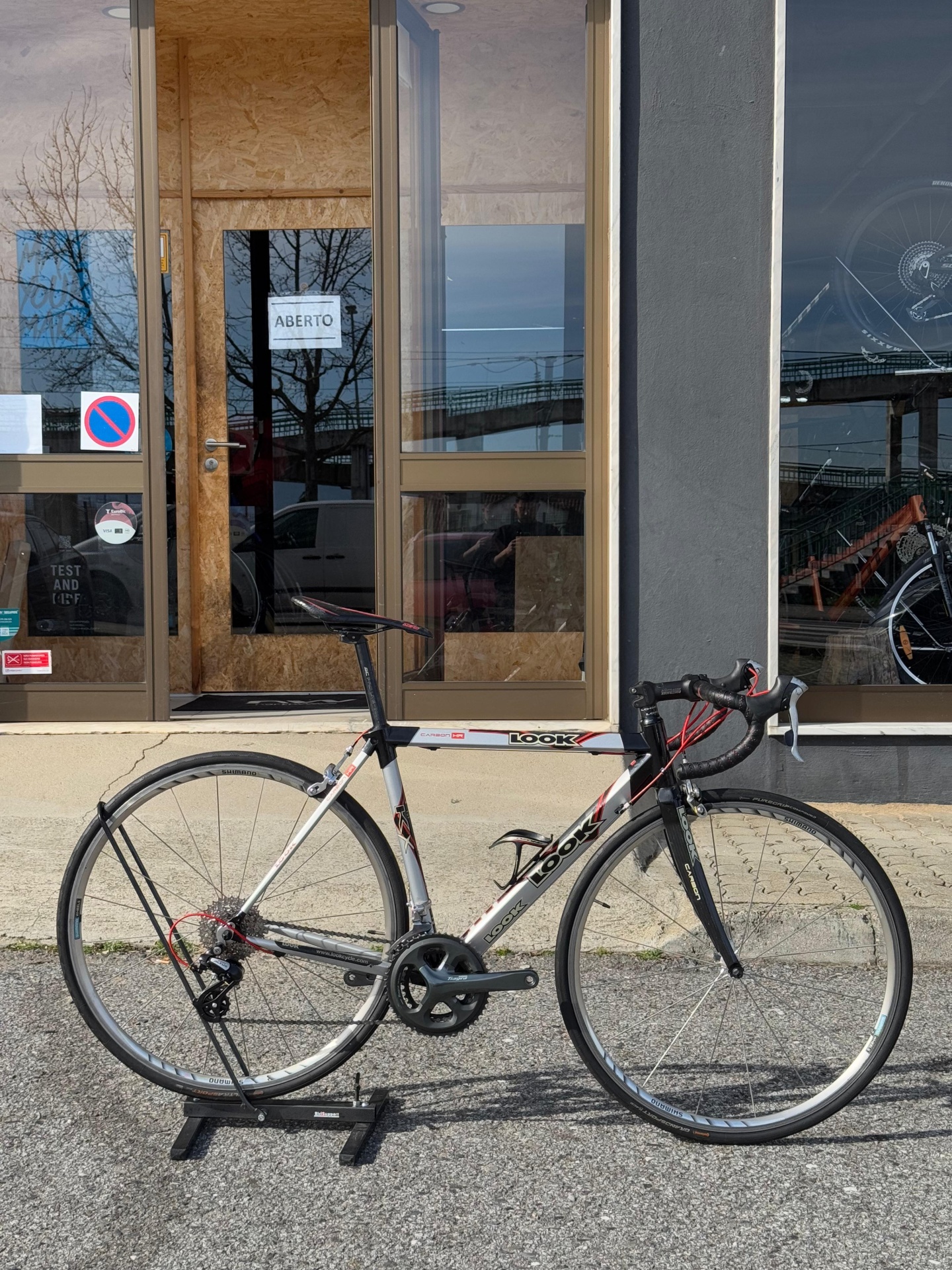 Bicicleta de estrada LOOK cinza, preto e vermelho em suporte diante de porta com placa ABERTO.