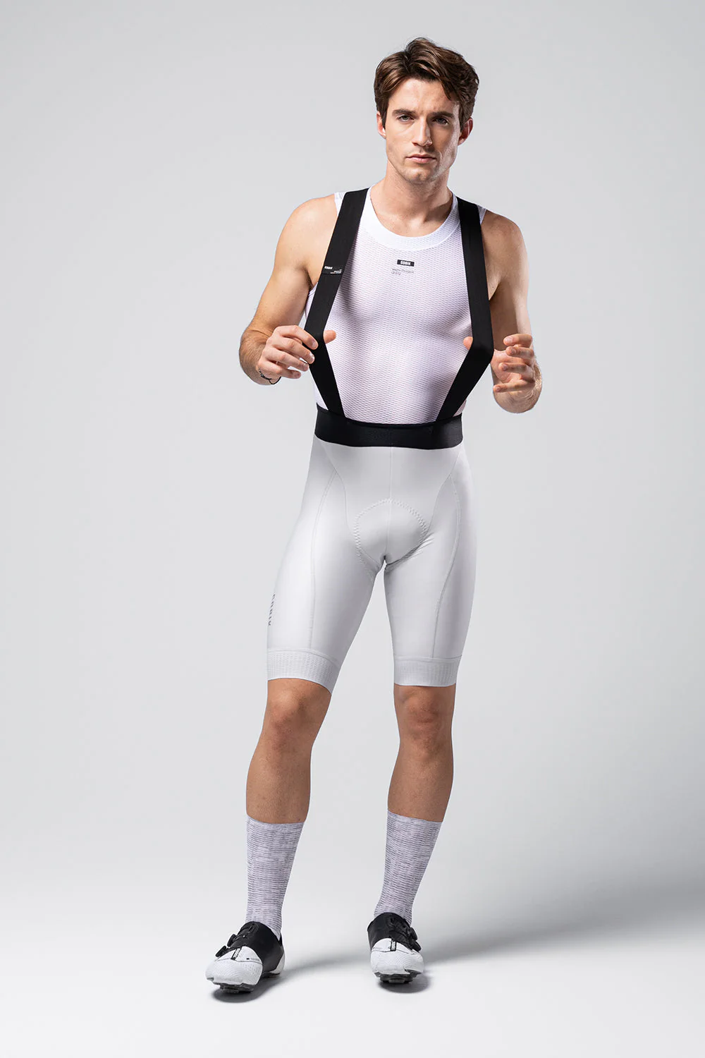 Homem com roupa desportiva branca e suspensórios pretos num fundo cinza claro.