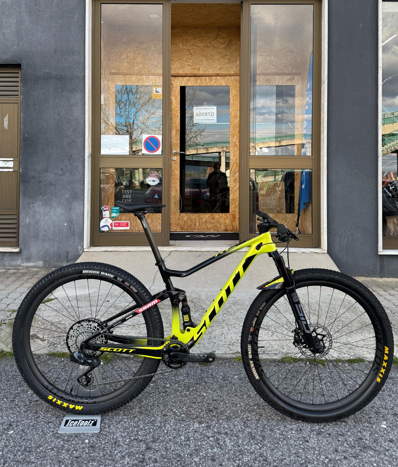 Bicicleta Scott preto e amarelo encostada em parede cinzenta frente à porta de madeira