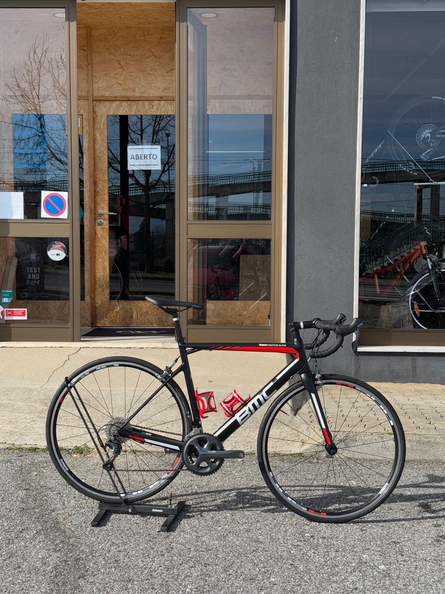 Bicicleta de estrada BMC preta e vermelha junto a porta de vidro com letreiro ABIERTO