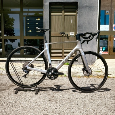 Bicicleta de estrada cinzenta com roda fina e guidão preto apoiada num suporte à frente de edifício.