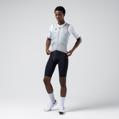 Homem com colete cinzento, camiseta branca, calções de ciclismo pretos e sapatos brancos em fundo cinzento