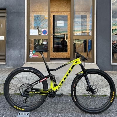 Bicicleta Scott preto e amarelo encostada em parede cinzenta frente à porta de madeira