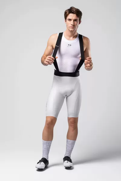 Homem com roupa desportiva branca e suspensórios pretos num fundo cinza claro.