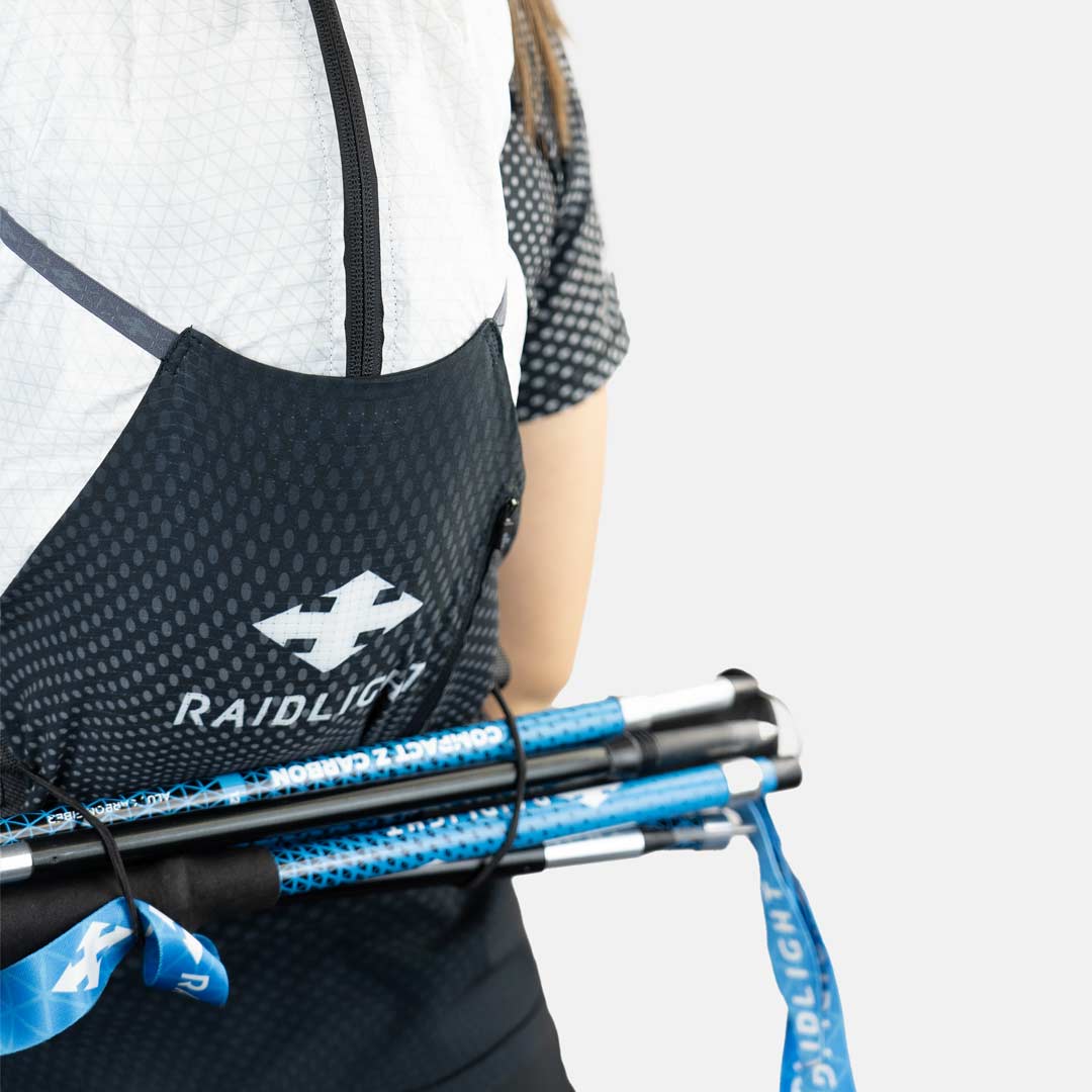 Pessoa com equipamento Raidlight preto e branco segurando bastões de caminhada azuis