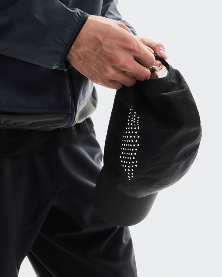 Pessoa segurando boné preto com perfurações circulares, veste casaco e calças pretas