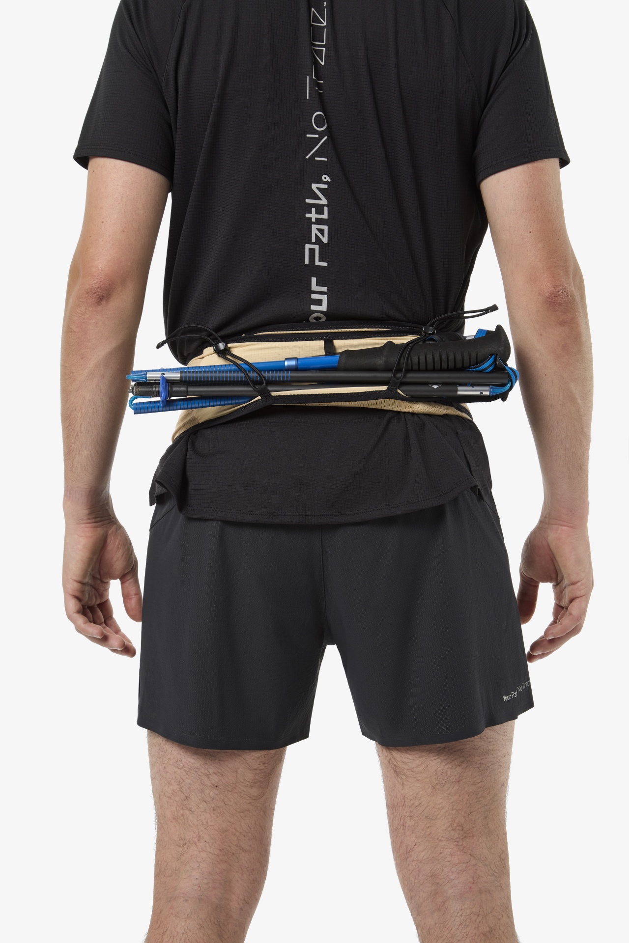 Pessoa de costas com camisola preta com texto branco, calções pretos e cinto bege com bastões de trekking azuis e pretos na parte traseira