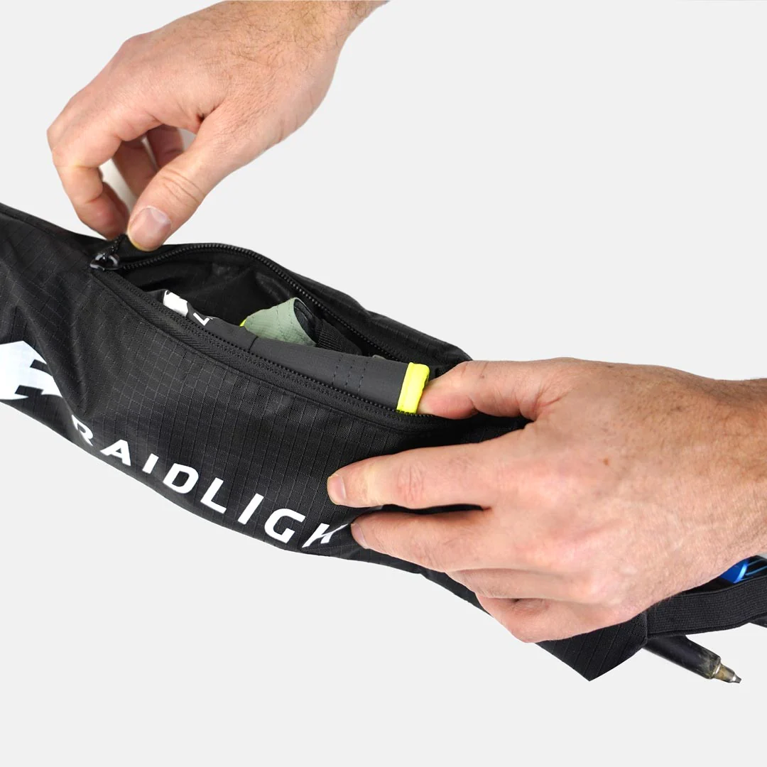 Bolsa preta com fecho e texto RAIDLIG, mãos a abri-la mostrando interior com objeto cinzento e amarelo