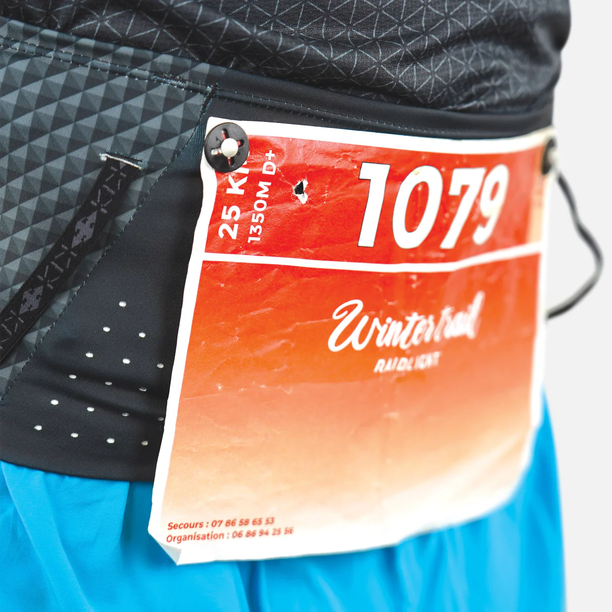 Etiqueta de corrida vermelha com número 1079 presa à roupa preta com padrão geométrico e calções azuis