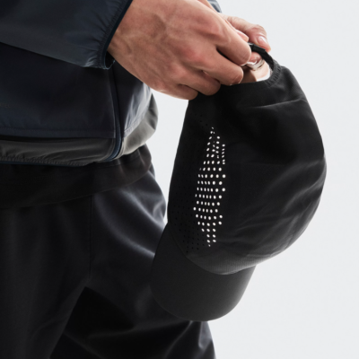 Pessoa segurando boné preto com perfurações circulares, veste casaco e calças pretas