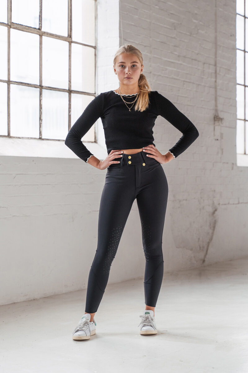 Mulher com calças justas pretas, camisola preta e sapatilhas brancas num estúdio minimalista