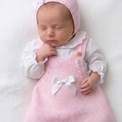 Bebé vestida com vestido rosa de malha, camisola branca e gorro rosa com laço branco, deitada em superfície branca