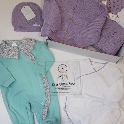 Conjunto de roupa de bebé em verde água, lavanda e branco sobre superfície branca.