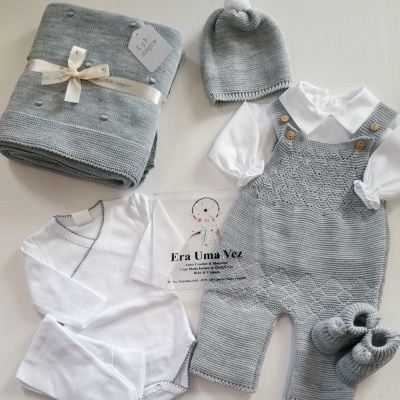 Conjunto de roupa de bebê com várias peças em branco e cinza, incluindo jardineira, gorro, body, manta e sapatinhos.