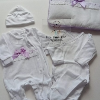 Conjunto de roupa de bebé branco com fita roxa e renda, e cartão de visita da loja Era Uma Vez