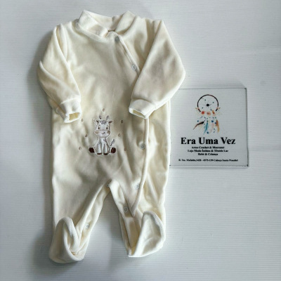 Babygrow creme com bordado de girafa sobre superfície branca com placa de loja ao lado.