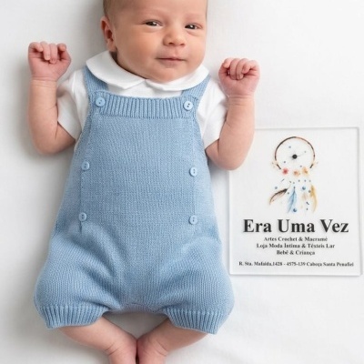 Bebé com macacão azul claro e camisola branca a sorrir deitado junto a cartaz com texto informativo.