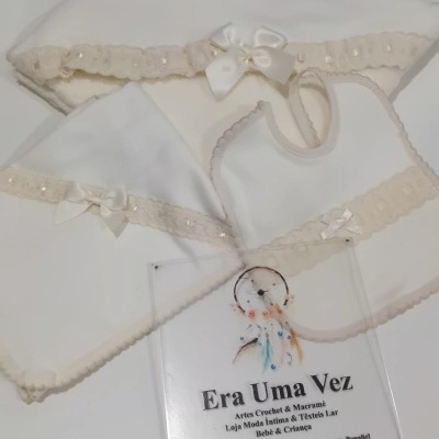 Conjunto de roupa de bebé branca com bordado bege, com placa informativa de loja sobre fundo branco.