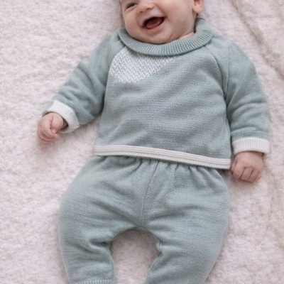 Bebé sorridente vestido com conjunto de malha verde água e branco deitado num cobertor branco.