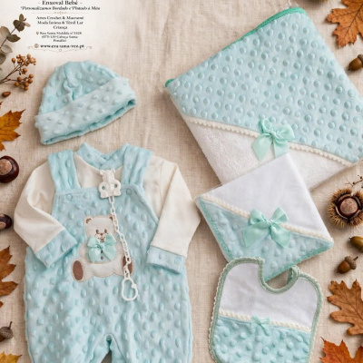 Conjunto para bebé azul claro com detalhes em relevo e laços, sobre tecido bege com folhas e bolotas ao redor.