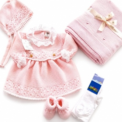 Conjunto de roupa de bebê cor-de-rosa com vestido, touca, sapatinhos, manta e meias sobre fundo branco.