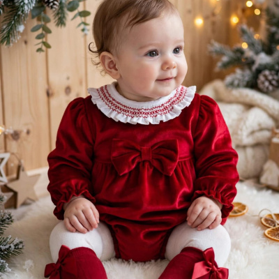 Bebé com roupa de Natal vermelha sentada em textura felpuda branca com decoração de pinhas e luzes.
