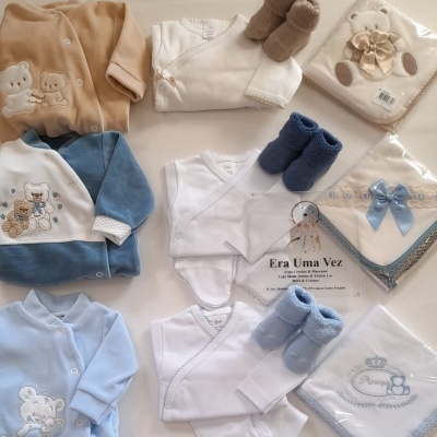Roupa de bebé com bodies, casacos, botinhas e panos bordados com ursinhos, em superfície clara.