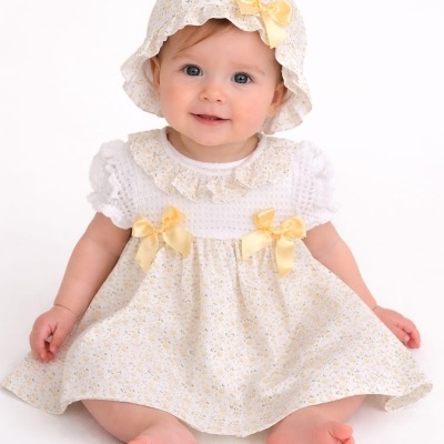 Bebé com chapéu e vestido floral amarelo sentado em fundo branco