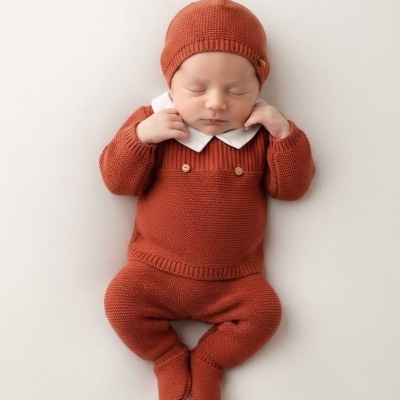Bebé com roupa de malha terra cota e gorro a dormir num fundo claro.