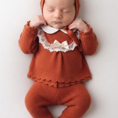 Bebé vestido com roupa de malha cor ferrugem, incluindo camisola com gola branca e touca, deitado sobre fundo branco.
