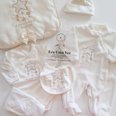 Conjunto de roupa de bebé branco com bordado estrela e texto, incluindo body, babete, macacão, gorro e manta com laço.