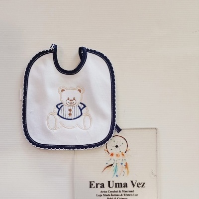 Babete branco com borda azul escura e ursinho bordado ao lado de cartão com texto Era Uma Vez