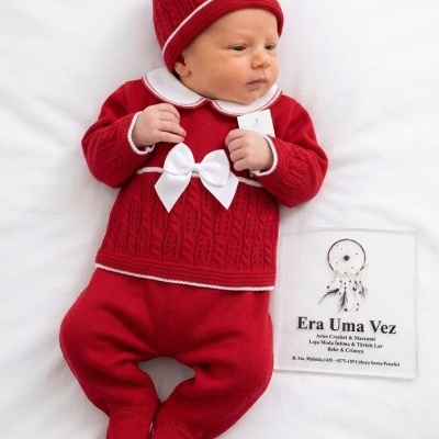 Bebé deitado com conjunto vermelho de malha e laço branco ao lado de papel com texto Era Uma Vez