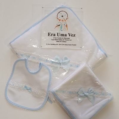 Conjunto de bébé branco e azul com babete, toalha e fralda decorados com laços em embalagem transparente.