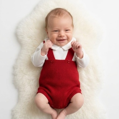 Bebé sorridente de body branco e jardineira vermelha, deitado numa pele branca.