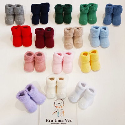 Vários pares de botins para bebé coloridos dispostos em fila com cartão informativo em fundo branco.