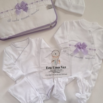 Conjunto de roupa de bebé branco com detalhes lilás e saco bordado com frase em português.