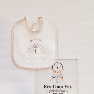 Babete branco com bordado de ursinho sobre superfície branca com placa decorativa e texto