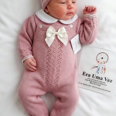 Fato de bebé rosa com gola branca e laço, gorro da mesma malha, deitado em cama branca ao lado de saco era uma vez