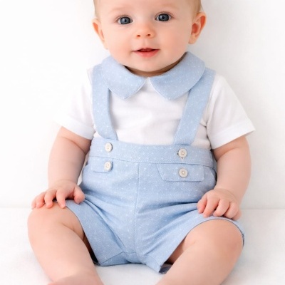 Bebé de olhos azuis vestindo calções azuis com suspensórios e body branco sentando-se