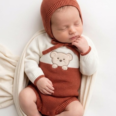 Bebé com macacão e gorro de malha castanho e branco a dormir numa manta creme