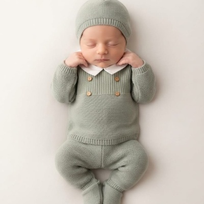 Bebé vestido com roupa de malha cinza-verde clara e gorro, em fundo branco.