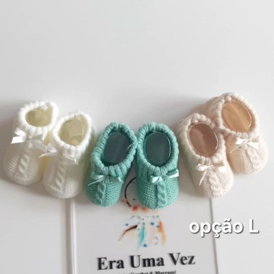 Sapatinhos de bebê de crochet em branco, verde e bege com laços, sobre superfície branca com cartão informativo